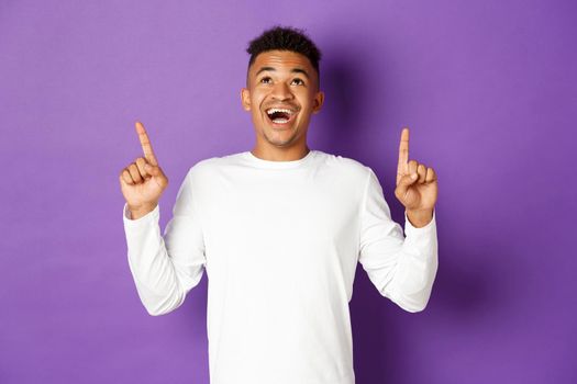 Handsome excited african-american guy, wearing white sweatshirt, looking amazed and pointing fingers up, showing super cool advertisement, standing over purple background.