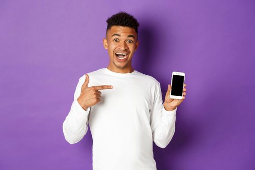 Handsome and excited african-american guy, pointing finger at smartphone screen, showing shopping site or app, standing over purple background.