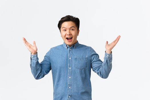 Happy and excited asian surprised guy receive awesome news, raising hands sideways and smiling amazed, praising great job, saying congrats, rejoicing over white background.