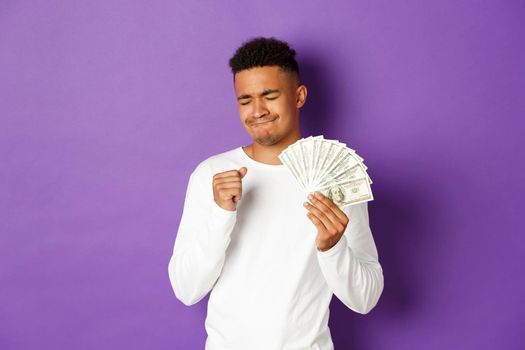 Image of pleased african-american man, feeling satisfaction of winning money, holding cash and rejoicing, standing over purple background.