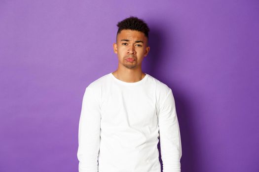 Portrait if silly african-american guy whining, begging for something, crying and looking sad at camera, standing over purple background.