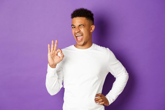 Image of handsome african-american guy, winking cheeky and smiling, showing okay sign, approve something, being satisfied, standing over purple background.
