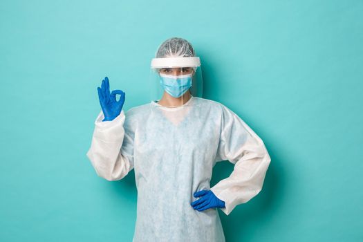 Concept of covid-19, pandemic and health. Female doctor in personal protective equipment, looking confident and determined, showing okay gesture, standing over blue background.