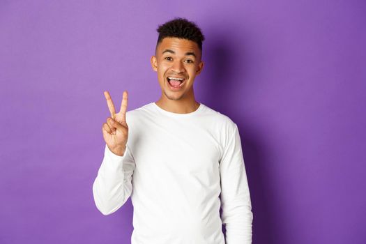 Image of cheerful african-american guy in white sweatshirt, showing two fingers and smiling, standing over purple background.