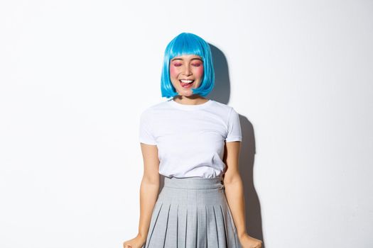 Image of cheerful asian girl in blue wig smiling carefree, close eyes and daydreaming, standing over white background.