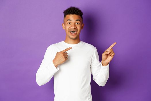 Image of happy african-american male model in white sweatshirt, pointing fingers at upper right corner and smiling, showing promo, standing over purple background.