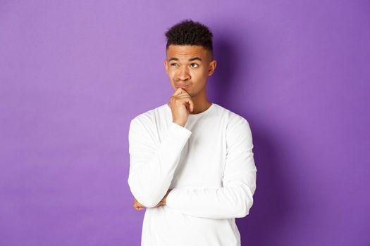 Portrait of young african-american male student thinking, looking at upper left corner thoughtful, making choice, standing over purple background with copy space.