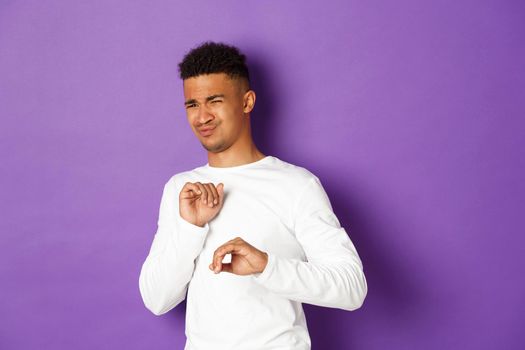 Image of young african-american guy cringe and looking at something disgusting, grimacing and standing over purple background.