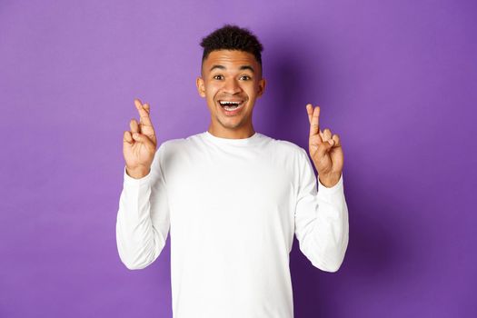 Image of handsome african-american young guy in white sweatshirt, looking hopeful and smiling with crossed fingers, making wish or waiting for good news, standing over purple background.