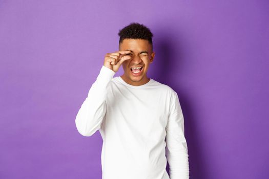Happy african-american man showing something small, promote tiny thing, standing over purple background in white sweatshirt.