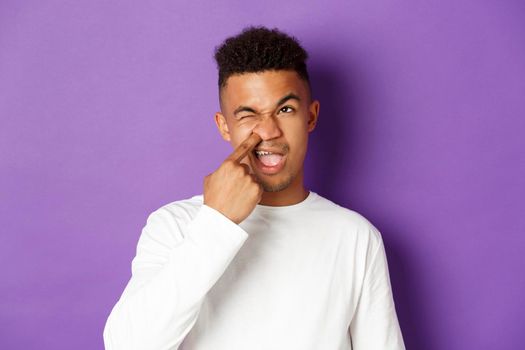 Image of young african-american man picking nose and looking upper left corner spaced out, standing over purple background.