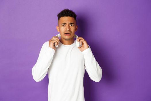 Image of modern african-american man, take-off headphones and looking confused, standing over purple background.