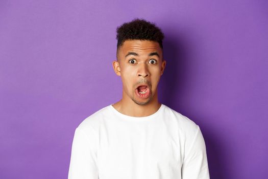 Close-up of impressed african-american man, drop jaw and stare at something amazed, standing over purple background.