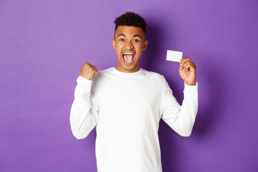 Portrait of handsome african-american man, looking excited and cheerful, showing credit card and making fist pump gesture, standing over purple background.