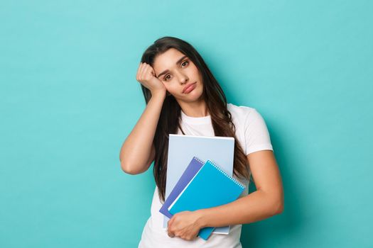 Portrait of tired pretty female student, holding study material and notebooks and looking exhausted, standing over blue background.