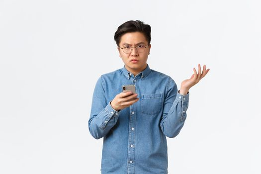 Confused and disappointed asian guy in glasses cant understand reasons, standing white background, raising hand up puzzled after seeing something frustrating in mobile phone, white background.