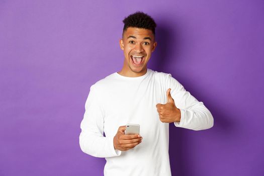 Image of amazed and satisfied african-american guy, holding mobile phone and showing thumbs-up in approval, like something, standing over purple background.
