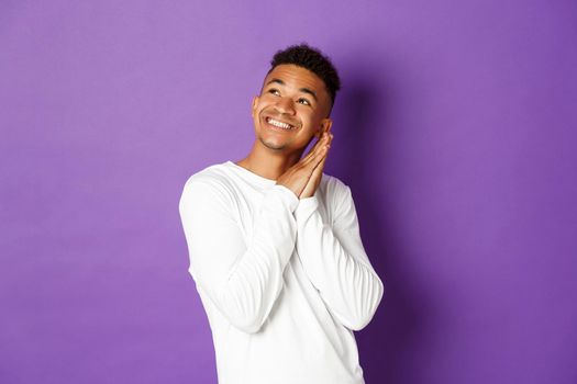 Image of happy african-american man in white sweatshirt, feeling pleased, looking dreamy at upper left corner and smiling, imaging something, standing over purple background.