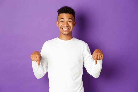Portrait of african-american happy guy, wearing white sweatshirt, pointing fingers down and smiling cheerful, showing advertisement, standing over purple background.