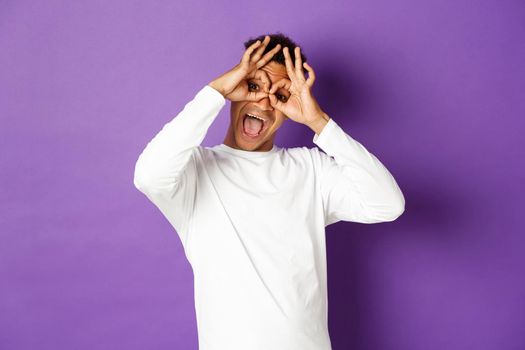 Image of funny and happy african-american man in white sweatshirt, grimacing and making faces, standing over purple background.