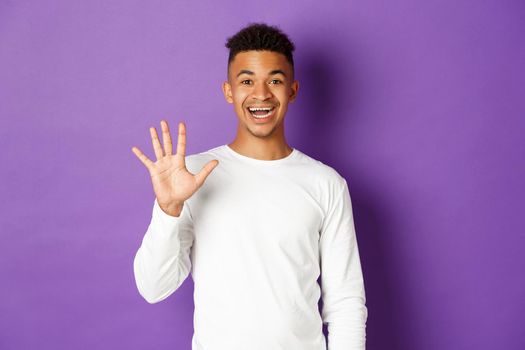 Image of cheerful african-american guy in white sweatshirt, showing number five and smiling, standing over purple background.