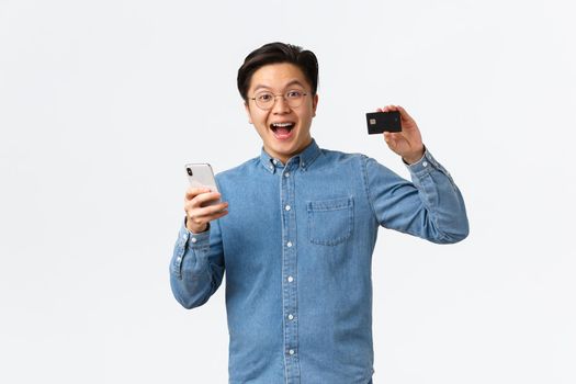 Excited and satisfied asian guy in glasses and casual clothes showing credit card with proud expression, paying for something in internet using smartphone, shopping online, white background.
