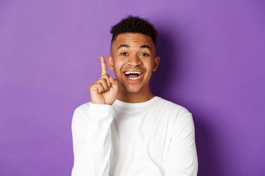 Image of thoughtful african-american man have an idea, raising finger and give suggestion, standing in white sweatshirt over purple background.