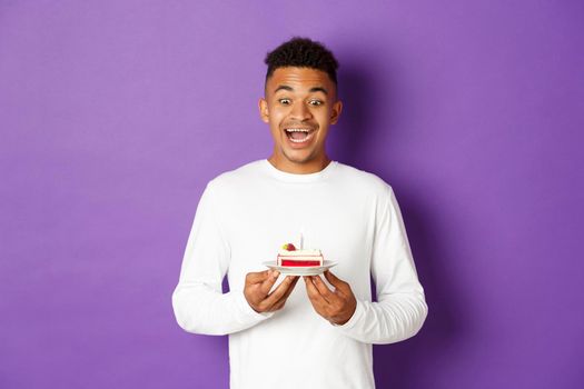 Image of happy african-american guy celebrating birthday, looking excited at b-day cake and making wish, standing over purple background.