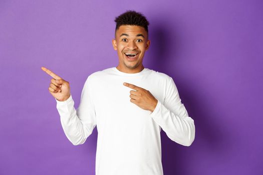 Image of excited and happy african-american guy pointing fingers at upper left corner, smiling amazed, showing cool advertisement, standing over purple background.