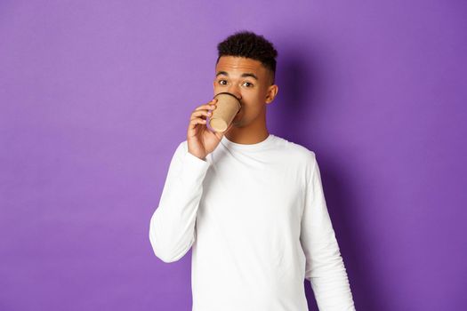 Image of handsome african-american male model, standing over purple background and drinking takeaway coffee.