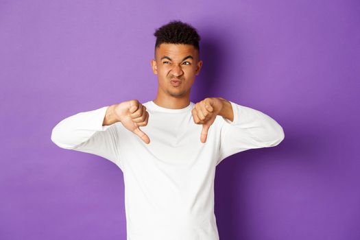 Image of skeptical and disappointed african-american young man, looking at something disgusting at upper left corner, showing thumbs-down and grimacing from dislike, standing over purple background.