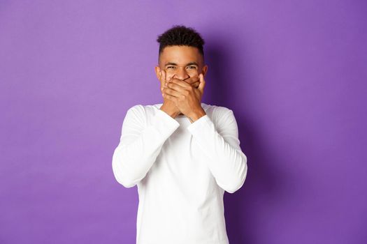 Image of shocked and disgusted african-american man, cover mouth, refrain from vomiting, standing over purple background.