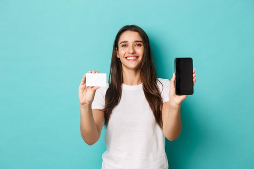 Portrait of happy attractive female model, showing mobile phone screen and credit card, smiling pleased, standing against blue background.