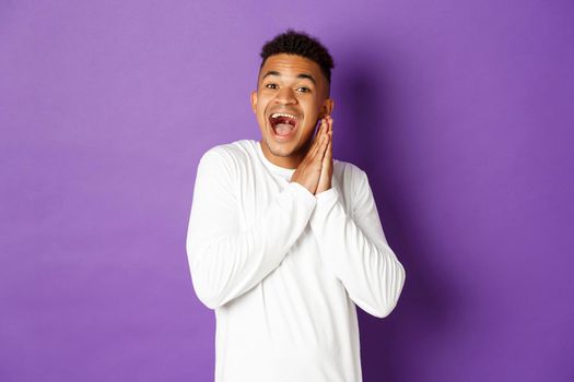 Image of excited and hopeful african-american queer guy, looking with anticipation and thrill at something, clap hands and smiling happy, standing over purple background.