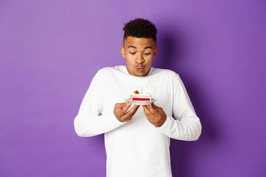 Happy african-american man celebrating birthday, blowing candle on a cake and making wish, standing over purple background.