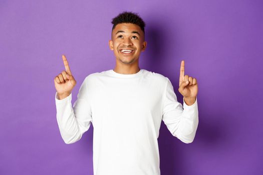 Cheerful african-american man in white sweatshirt, showing logo, pointing and looking up with happy smile, standing over purple background.