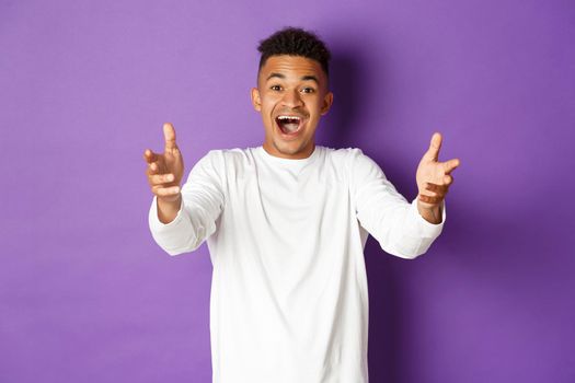 Portrait of handsome african-american young man, reaching hands forward to receive something, looking amazed and happy, standing over purple background.