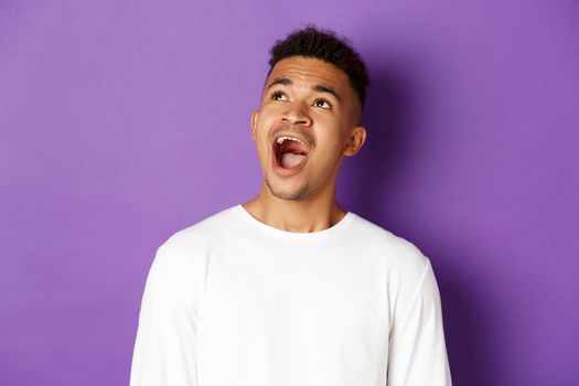 Close-up of amazed african-american young man, open mouth and saying wow, looking fascinated at upper left corner, standing over purple background with copy space.