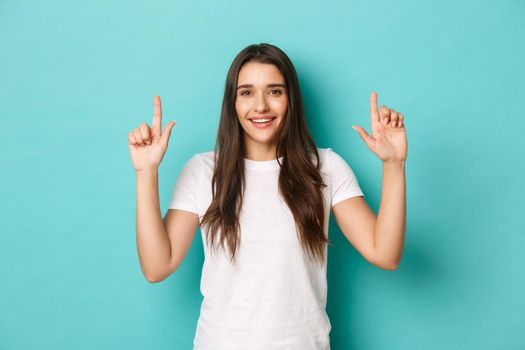 Portrait of happy beautiful woman in white t-shirt, smiling and pointing fingers up, showing logo, standing over blue background.