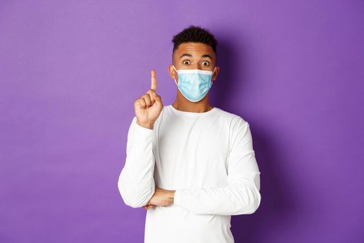 Concept of coronavirus, quarantine and lifestyle. Image of young african-american male in medical mask, raising finger up and suggesting plan, have an idea, standing over purple background.
