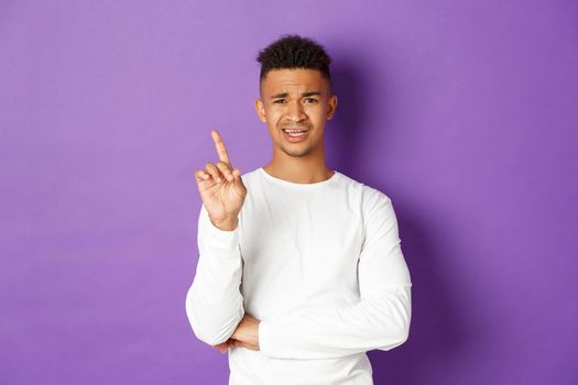 Image of skeptical young african-american man in white sweatshirt, forbid something, frowning disappointed and shaking finger in disapproval, dislike idea, standing over purple background.