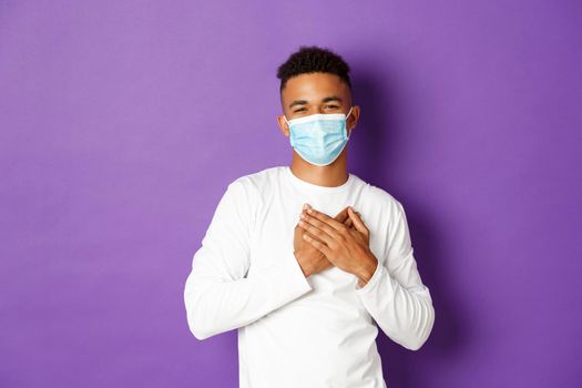 Concept of coronavirus, quarantine and lifestyle. Image of handsome african-american man in medical mask, saying thank you, standing grateful with hands on heart, standing over purple background.