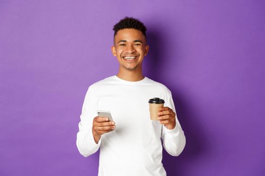 Portrait of handsome african-american guy smiling pleased, drinking coffee and surfing the net in mobile phone, standing over purple background.