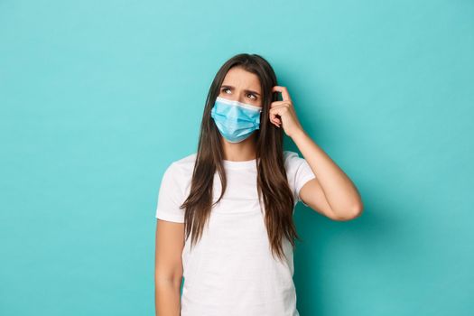 Concept of pandemic, covid-19 and social distancing. Image of puzzled young woman in white t-shirt and medical mask, looking confused at upper left corner, scratching head, blue background.