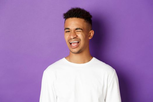 Close-up of handsome young african-american man winking, smiling and looking cheerful, standing over purple background.