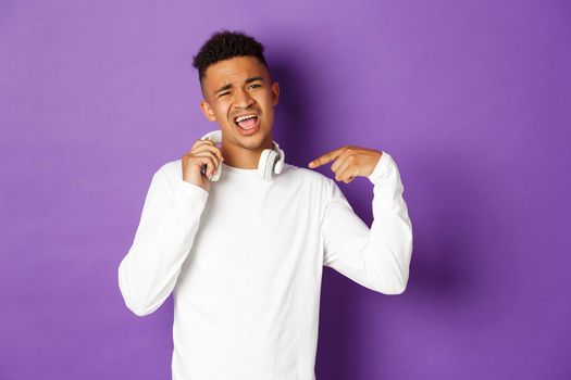 Image of sassy african-american guy, pointing at headphones and praising awesome song, standing over purple background.