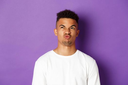 Close-up of troubled young african-american guy, looking up and grimacing perplexed, having hard choice, standing over purple background.