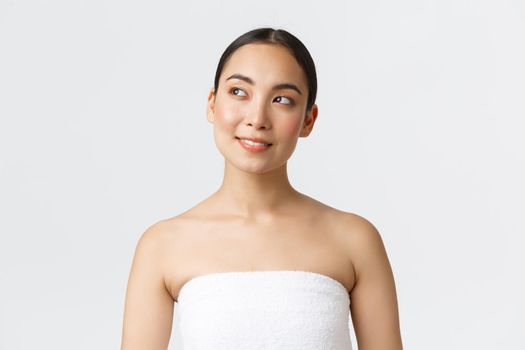 Thoughtful beautiful asian woman with perfect clean skin standing in towel and looking curious upper left corner, thinking, see skincare or haircare product advertisement, visit spa salon.