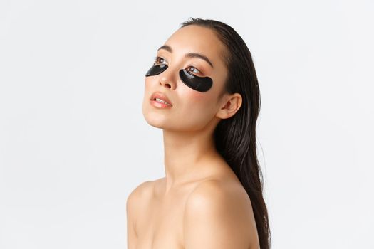 Skincare, women beauty, hygiene and personal care concept. Profile of attractive sensual asian woman standing naked and looking away, touching clean skin, using eye patches, remove dark circles.