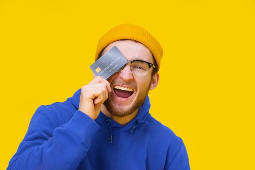 Happy young caucasian guy with credit, debit card in hand smiling cover his eye ready to spend money. Young man in blue hoodie with mockup bank card isolated on yellow background. Shopping concept.